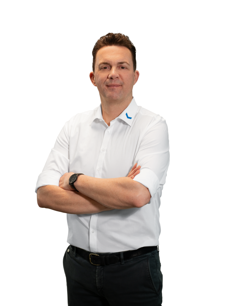 A man in a white shirt with his arms crossed, standing in front of a neutral background.